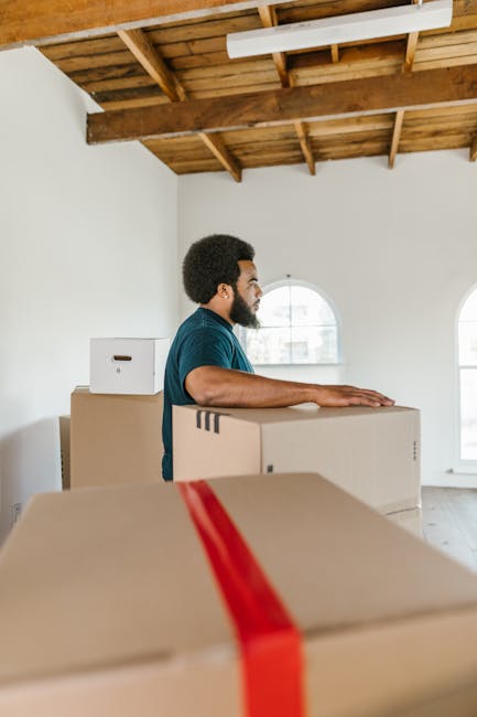A man with dark skin, curly hair, and a beard is standing inside a spacious room with a high wooden ceiling and large arched windows, holding a large cardboard moving box. Several other cardboard boxes of varying sizes, some sealed with red packing tape, are stacked around him, indicating a packing and loading process during a home relocation. The room has white walls and natural daylight streaming through the windows. In the background, there is a partially visible white wall and the ceiling structure made of exposed wooden beams. The scene depicts a professional moving operation, with [COMPANY_NAME], such as Stockwell Removals, assisting with furniture transport and packing services, focusing on the efficient and careful handling of household belongings during a same-day SW9 removals service.
